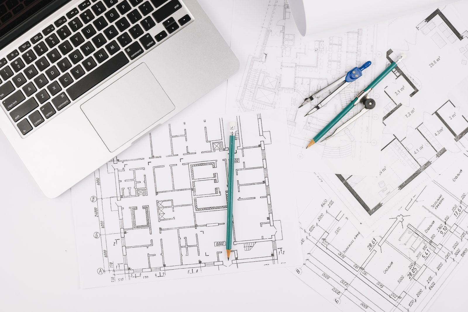 Laptop beside printed floor plan drawings with a drafting compass and pencil