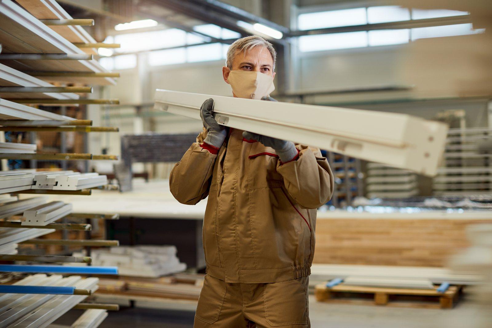 Worker in a protective mask carrying a cut panel through a carpentry workshop