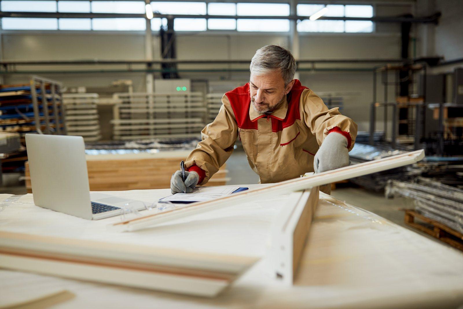 Worker noting measurements against a clipboard beside cut boards and a laptop in a workshop